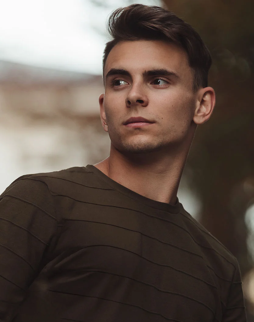Close-up of a young man with short, dark brown hair styled upwards. He has a neutral expression, focused eyes, and wears a dark olive green long-sleeved t-shirt with a ribbed texture. The blurred background hints at an urban or outdoor setting, creating a casual portrait vibe. - Earlobe Reduction in Glenview, IL