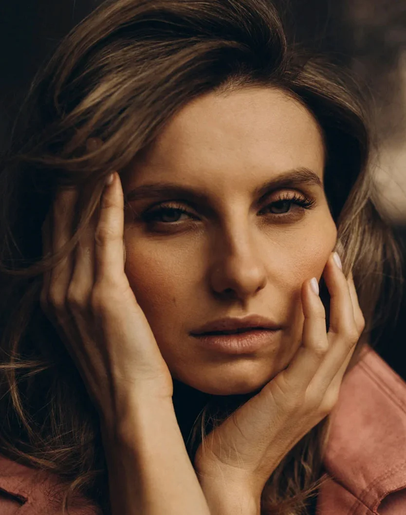 Close-up of a woman with medium-length wavy brown hair, a contemplative expression, and eyes looking directly at the viewer. Her hands gently rest on her face, one on her forehead and the other on her cheek. She has smooth, light-tan skin and well-defined features. The dark background and soft lighting highlight her face, with a dusty rose jacket visible at the edges. - Facial Fat Grafting in Glenview, IL