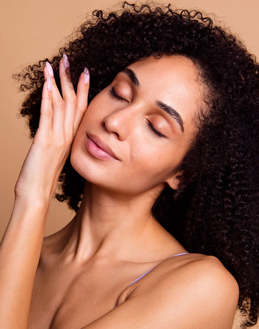 Close-up of a light-brown-skinned woman with curly dark hair, eyes closed and a serene expression. She wears a light lavender top, and soft lighting highlights her smooth skin and natural features against a light beige background. - Facial Paralysis in Glenview, IL