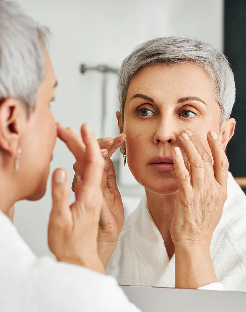 Close-up of a middle-aged woman with short gray hair examining her face in a mirror, touching her cheeks and eye area with a thoughtful expression. Soft lighting highlights her skin's natural texture and fine lines. She wears a white bathrobe, and the mirror's reflection clearly captures her face and hands. The background is blurred, keeping the focus on her and her reflection. - Skin Cancer Reconstruction in Glenview, IL