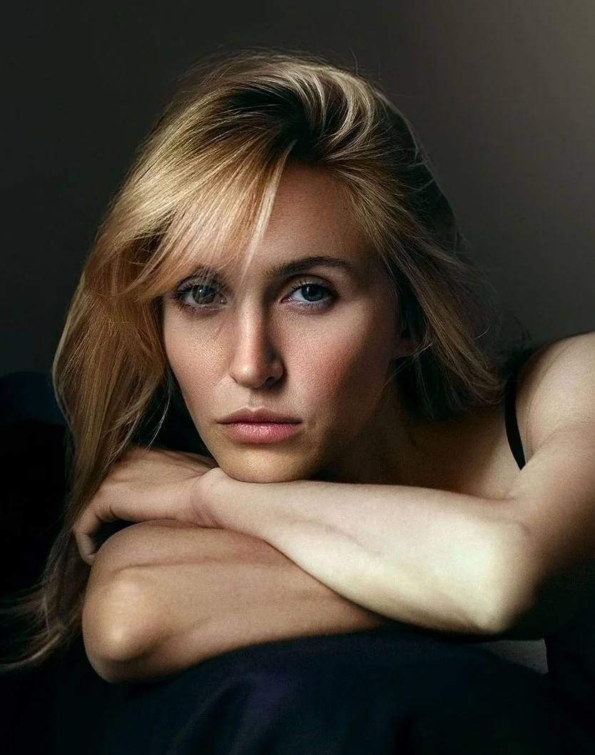 Close-up of a blonde woman resting her head and elbows on a surface, gazing downward with a contemplative expression. Soft lighting highlights her features, while a dark background enhances focus on her face. - Upper Eyelid Surgery in Glenview, IL
